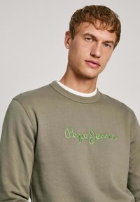 Sudadera verde oliva de cuello redondo, con el logo bordado en verde "Pepe Jeans" en el pecho. Tejido suave y corte holgado.