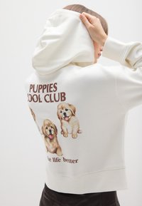 Crème-kleurige hoodie met een zachte textuur. Voorzien van een grafisch ontwerp van vier puppy's en de tekst "PUPPIES COOL CLUB" in bruine lettertype op de achterkant.