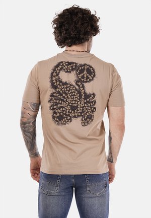 T-shirt corta beige a maniche corte con un grande design di uno scorpione nero stampato sul retro. Tessuto morbido, vestibilità standard. Abbinato a jeans blu.