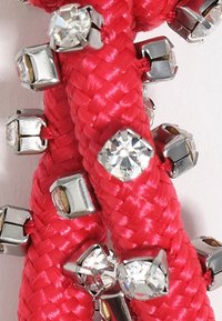 Svetlo roza pletena vrvica z metalnimi akcenti in vključenimi prozornimi rhinestones. Ima sijočo teksturo in angularno, geometrijsko zasnovo.