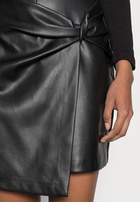 Jupe en cuir noir avec un détail noué à la taille, une texture douce et un ourlet anglé, offrant une silhouette ajustée.