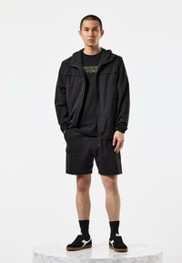 Black zip-up jacket with a hood, paired with a black t-shirt featuring gold text, black shorts, and black sneakers with white accents.