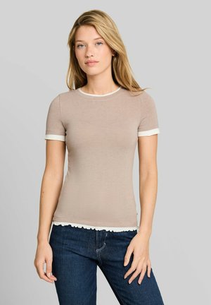 Giovane donna con lunghi capelli biondi che indossa una maglietta beige a maniche corte con bordo bianco e jeans blu scuro su sfondo grigio.