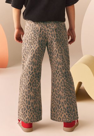 Barn iført vidtbenede leopardmønstrede bukser, sort trøje og røde sneakers, står mod en abstrakt beige og orange baggrund.