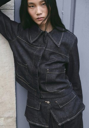 Persona joven con cabello largo y negro, vestida con chaqueta de mezclilla oscura y pantalones a juego, de pie contra una pared texturizada con una expresión neutra.