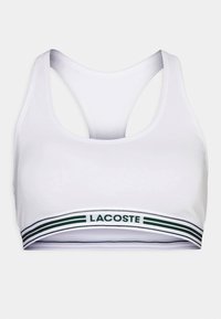 Biely športový podprsenka s pretekárskym strihom a zeleno-bielym pruhovaným pásom s logom "Lacoste". Hladká textúra látky.