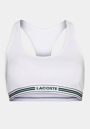 Biały biustonosz sportowy z wycięciem na plecach i zielono-białym paskiem z logo "Lacoste". Gładka struktura tkaniny.
