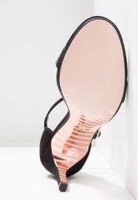 Chaussure à talon haut noire avec semelle réfléchissante en or rose, inclinée verticalement contre un mur d'angle blanc et une surface.