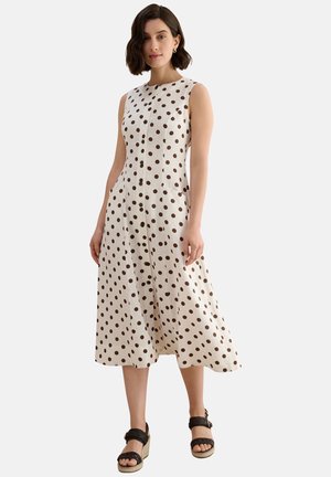 Mujer con vestido midi blanco sin mangas con lunares marrones y sandalias negras de cuña, de pie frente a un fondo liso.