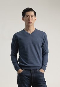 Suéter de punto azul marino con escote en v, confeccionado en una tela suave, que cuenta con mangas largas y un pequeño logo en el pecho, combinado con jeans oscuros.