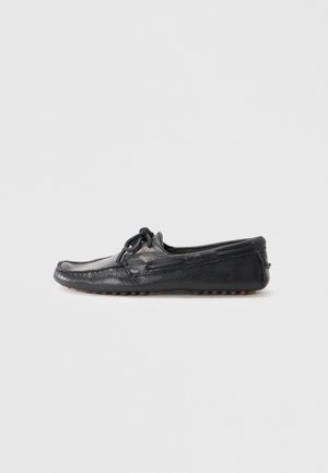 Sapato mocassim preto em couro, com bico redondo, detalhe de atacadores e sola de borracha com pinos para melhor aderência. Textura suave com costuras visíveis.