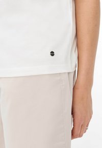 Hvid bomulds t-shirt med et rundt, sort logo patch nær sømmen, med en glat tekstur og en afslappet pasform, parret med lysebeige bukser.