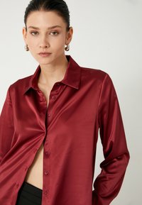 Camisa roja de seda con cuello y mangas largas. Presenta una tapeta delantera y una textura brillante, resaltando su acabado suave.