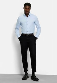 Hellblaues Hemd mit Knopfleiste, schwarze, maßgeschneiderte Hose, schwarzer Gürtel und Schuhe. Glatter Stoff, taillierte Form und lange Ärmel.