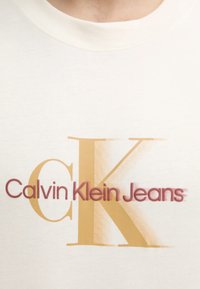 Camiseta de algodón crema con un gran logo gráfico en naranja y burdeos que dice "Calvin Klein Jeans" con un diseño de fuente distintivo.