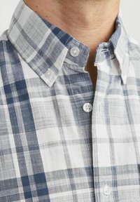 Camisa de botonadura en patrón de cuadros con tonos azules y blancos. Cuello con solapa, presenta botones de plástico blanco y una tela de algodón texturizada.
