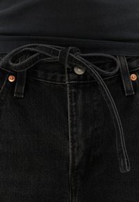Jean en denim noir avec rivets en cuivre et ceinture en tissu noir nouée à la taille, associé à un haut noir.