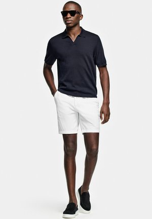 Man draagt een zwart poloshirt met korte mouwen, witte shorts, zwarte instapschoenen en een zonnebril, lopend met één hand in de zak op een witte achtergrond.