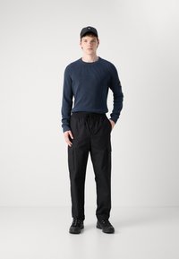 Maglione blu a trama, pantaloni cargo neri con tasche laterali, sneakers nere e un cappellino nero; outfit informale su uno sfondo semplice.