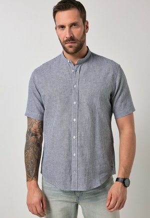 Mann trägt ein kurzärmeliges graues kariertes Hemd mit Knöpfen, helle Jeans, schwarze Armbanduhr und sichtbares Arm-Tattoo.