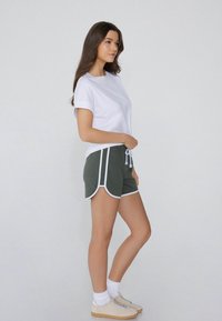 Witte T-shirt, groene shorts met witte bies en beige sneakers. De shorts hebben een trekkoord en een losse pasvorm.