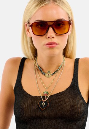Lunettes de soleil surdimensionnées marron, colliers superposés avec des accents dorés, motifs complexes et pierres précieuses colorées ; débardeur noir côtelé.