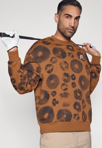 Καφέ φούτερ με κυκλικά μαύρα σχέδια, με στρογγυλή λαιμόκοψη και μανίκια με μανσέτες. Φοράει ανοιχτόχρωμο παντελόνι και κρατάει ένα μπαστούνι γκολφ.