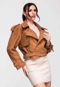 Femme portant une veste courte en daim marron avec des poignets à boucles et une robe en maille côtelée beige sur un fond uni.