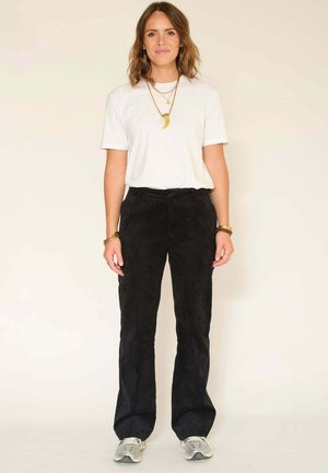 Pantalon en suède noir avec une coupe droite associé à un t-shirt blanc. Les accessoires incluent des colliers en or superposés. Chaussures de sport simples.