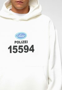 Witte hoodie met een voorzak, voorzien van een blauw ovale logo en een zwarte opdruk met de tekst "POLIZEI 15594" op de borst. Zacht, textuurachtig materiaal.