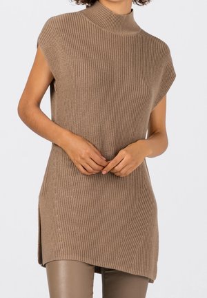 Brązowa dzianinowa tunika bez rękawów z wysokim kołnierzem i bocznymi rozcięciami, noszona na tle taupe skórzanych spodni, ręce złożone na talii.