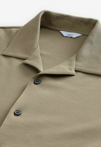 Beige Poloshirt aus glattem, strukturiertem Stoff. Hat einen breiten Kragen und zwei schwarze Knöpfe auf der Blende. Das Etikett trägt die Aufschrift "NEXT RELAXED FIT."