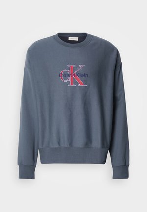 Szürke pamut pulóver kerek nyakkal, hosszú ujjakkal és bordázott mandzsettákkal, amelyen egy nagyméretű piros és kék "CK" logó található az elülső részén.