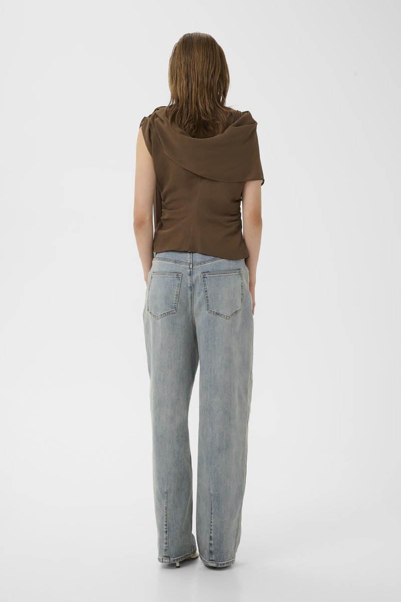 Femme debout, dos tourné, vêtue d'un haut marron sans manches et d'un jean taille haute bleu clair à jambes larges sur un fond uni.