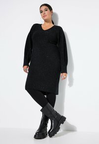 Schwarzes Strickkleid mit V-Ausschnitt, langen Ärmeln und Glitzerdetails. Kombiniert mit schwarzen Leggings und klobigen schwarzen Ankle Boots.