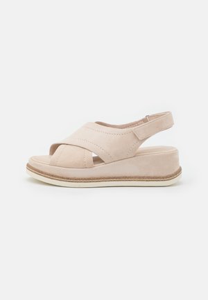 Beige suede sandal with cross-strap design, open toe, and adjustable heel strap. Features a light-colored platform sole with textured accents.