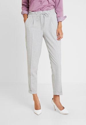 Pantalons tissés gris avec une coupe décontractée, taille élastique à cordon, et ourlets repliés ; associés à des chaussures à talons hauts blanches et une chemise violet clair.