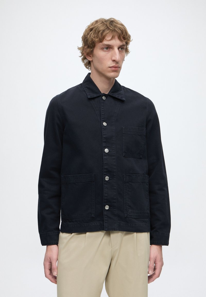 Jeune homme aux cheveux blonds bouclés portant une veste noire boutonnée avec trois poches avant et un pantalon beige sur un fond uni.