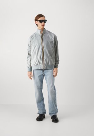 Giacca zip grigia chiara con colletto alto, polsini elastici e zip a contrasto; abbinata a jeans azzurri e scarpe nere.