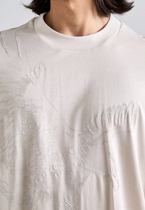 Jasnobeżowa koszula z teksturowanym wzorem ptaka na piersi, pokazana od szyi do połowy tułowia, z widocznymi krótkimi ciemnymi włosami.