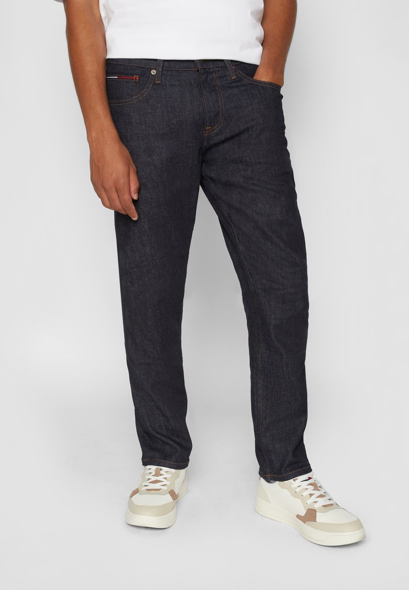 Donkerdenim jeans met een rechte pasvorm, voorzien van oranje stiksel en zakken. Gekleed met witte sneakers, die een gestructureerde stof tonen.