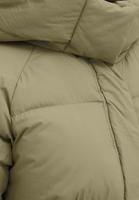 Puffjakke i en lys olivengrøn farve, med kviltede sektioner, glat tekstur og fremtrædende syninger.