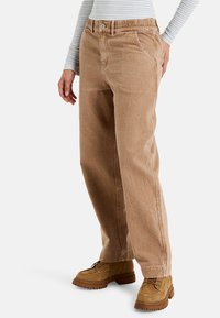 Pantalones de pierna ancha beige hechos de tejido texturizado con cierre de botón y cinturilla elástica; combinados con botas de tobillo marrón claro.