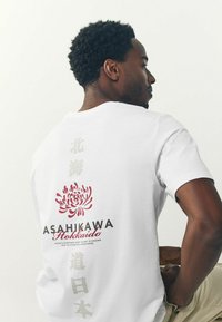 Hvid bomulds t-shirt med et blomstret grafisk design i rød og grå, som har teksten "ASAHIKAWA Hokkaido" på bagsiden og japanske tegn ovenover.