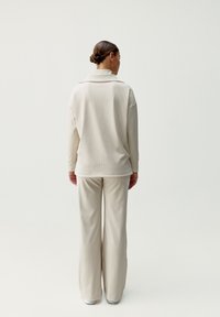 Krémszínű, hosszú ujjú blúz hajtókás gallérral, laza krémszínű nadrággal párosítva, lágy, minimalista megjelenést kölcsönöz. Sima szövettextúra.