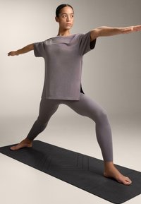 Šedý športový top s krátkymi rukávmi a bočnými rozparkami, v kombinácii s priliehavými legínami v rovnakom odtieni na čiernej jogovej podložke. Neutrálne pozadie.