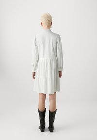 Lysegrønn, kragekjole med lagdelt skjørt, lange ermer, teksturert stoff og svarte cowboystøvler. Vist bakfra.