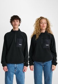 Sort fleece pullover med en kvart lynlås, der har en frontlomme med et logo. Begge modeller har afslappede denim jeans på.