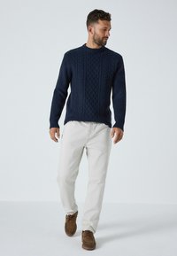 Marineblauer Kabelstrickpullover mit geripptem Muster, Rundhalsausschnitt, kombiniert mit hellen Hosen und braunen Freizeitschuhen. Model beim Gehen.