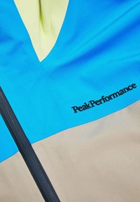 Blaue und beige Jacke mit einem V-förmigen Design, Reißverschluss und dem in Schwarz gestickten "PeakPerformance"-Logo. Glattes, leichtes Material.